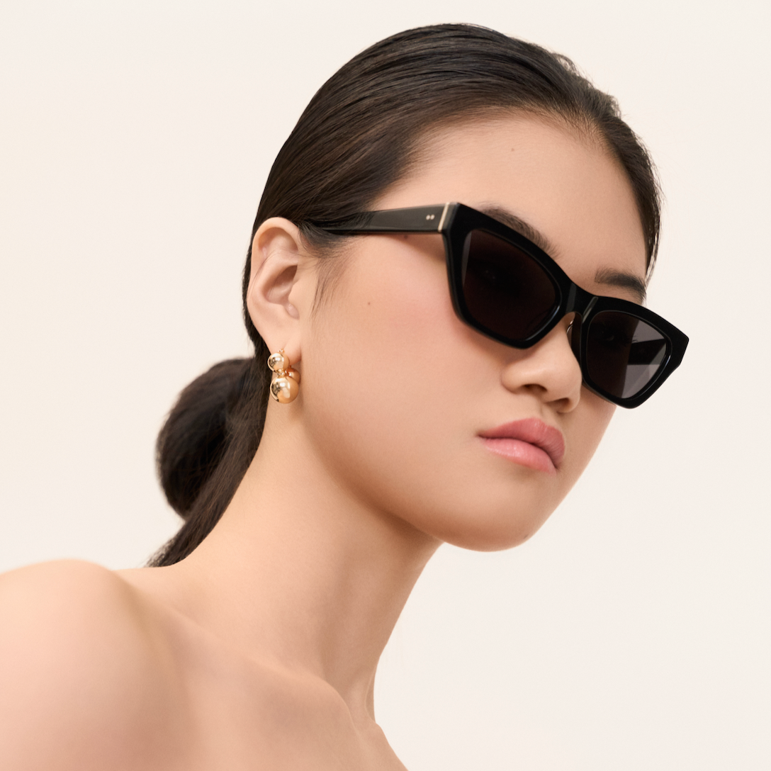 Woman wearing KayTran Eyewears Kai - Polished Black sunglasses, gold earrings, and sleek hair against a neutral background.