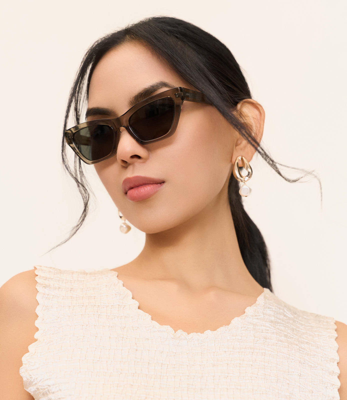 Woman wearing KayTran Eyewears Kai - Sage sunglasses, pearl earrings, and a textured sleeveless top.