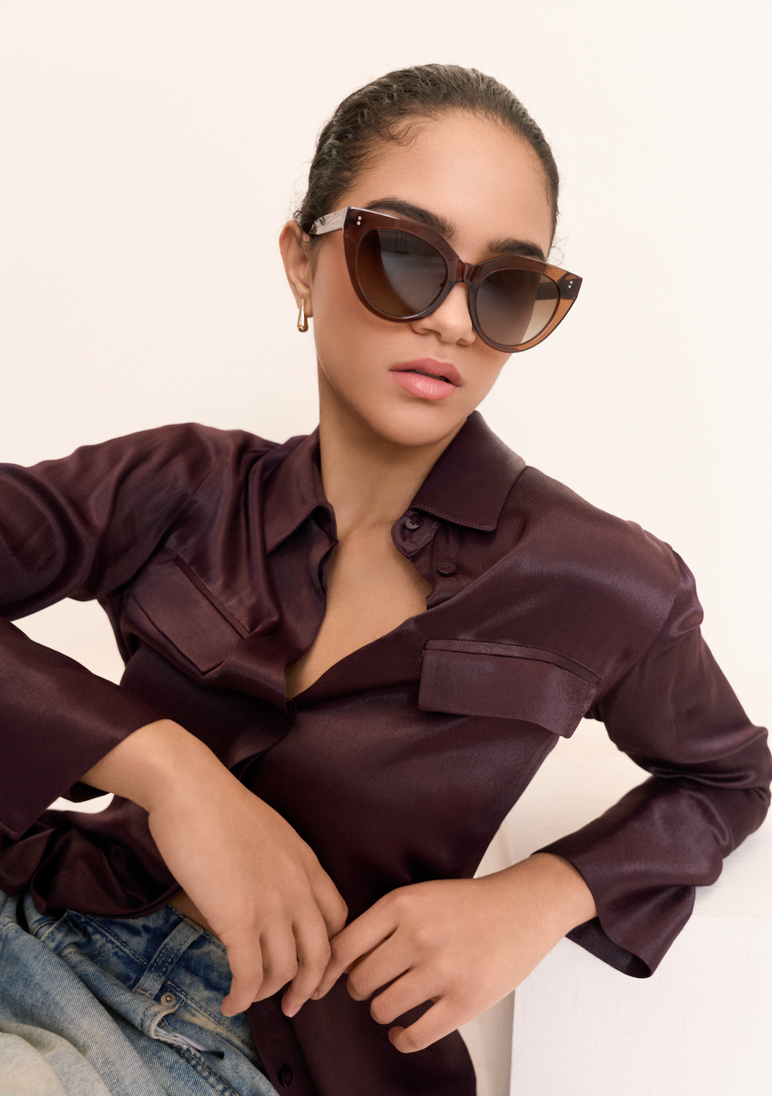 Woman wears kaytran eyewear Aiden - Truffle oversized sunglasses, a dark satin shirt, and jeans, seated by a white surface.