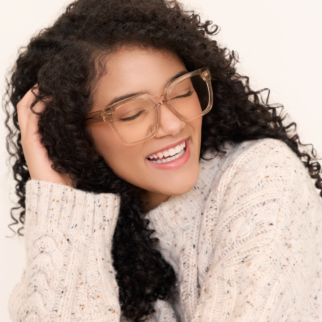 Woman with curly hair smiles with eyes closed, wearing KayTran Eyewear Andy - Folkstone Grey clear frames and a cream sweater.