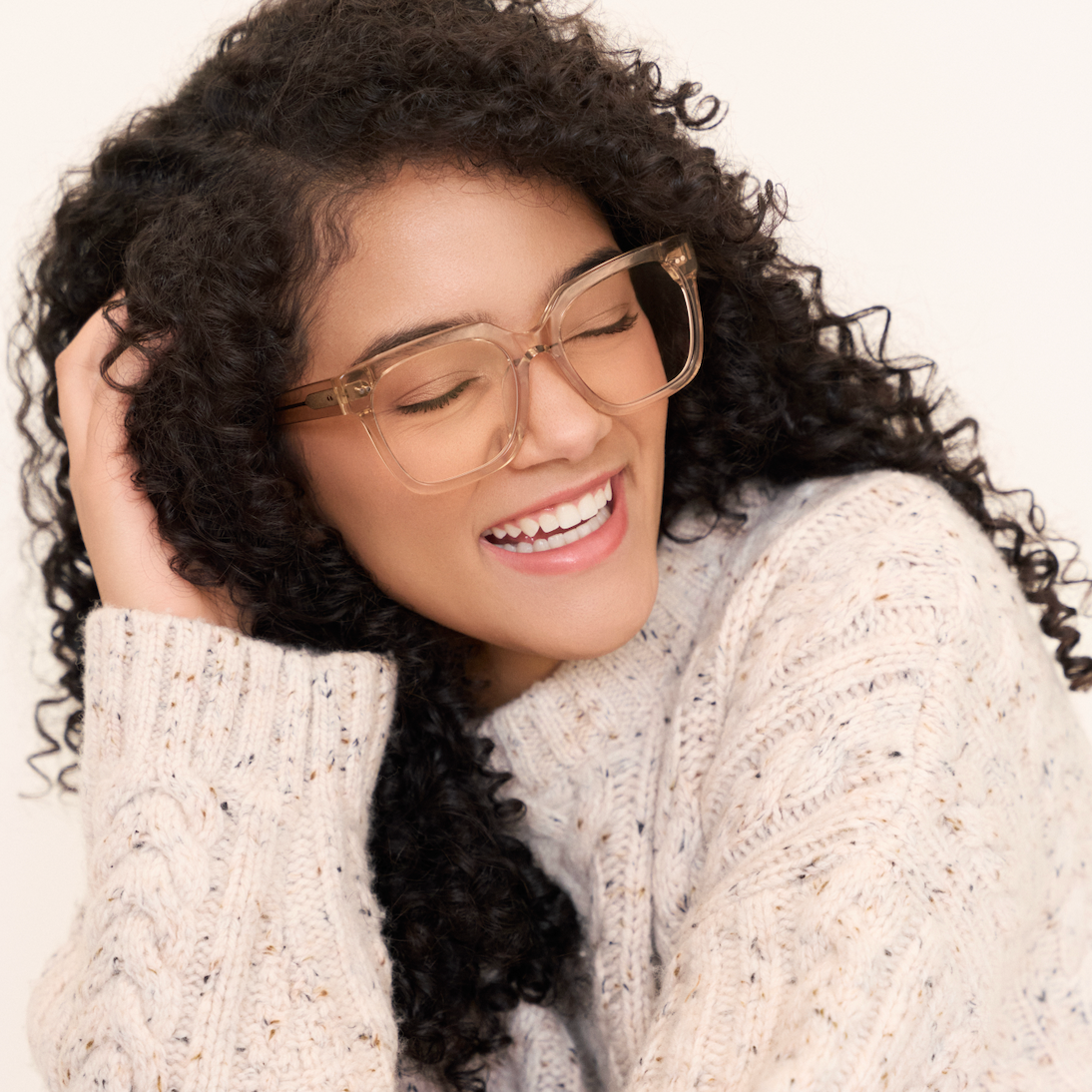 Woman with curly hair smiles with eyes closed, wearing KayTran Eyewear Andy - Folkstone Grey clear frames and a cream sweater.