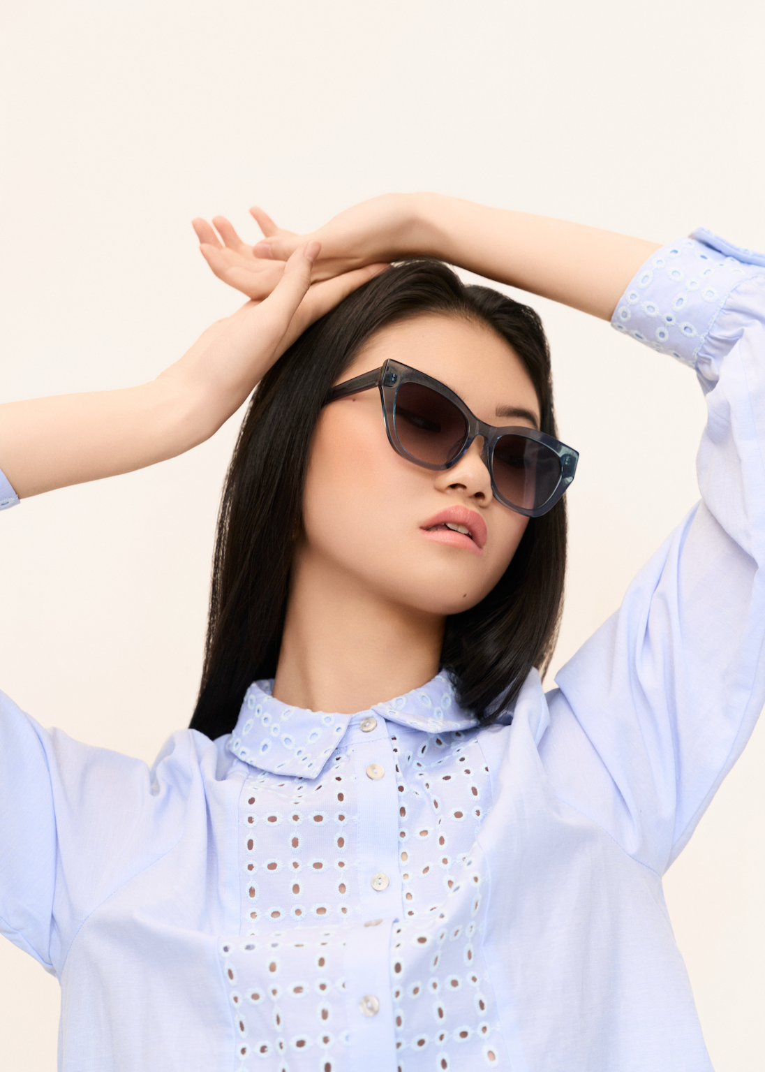 Woman wears KayTran Eyewears Harper - Sea Pine and a light blue blouse, posing with arms raised against a plain background.