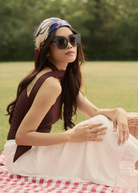 A woman in KayTran Eyewears Rowan - Sage sunglasses and a headscarf sits outdoors on a picnic blanket amid greenery.