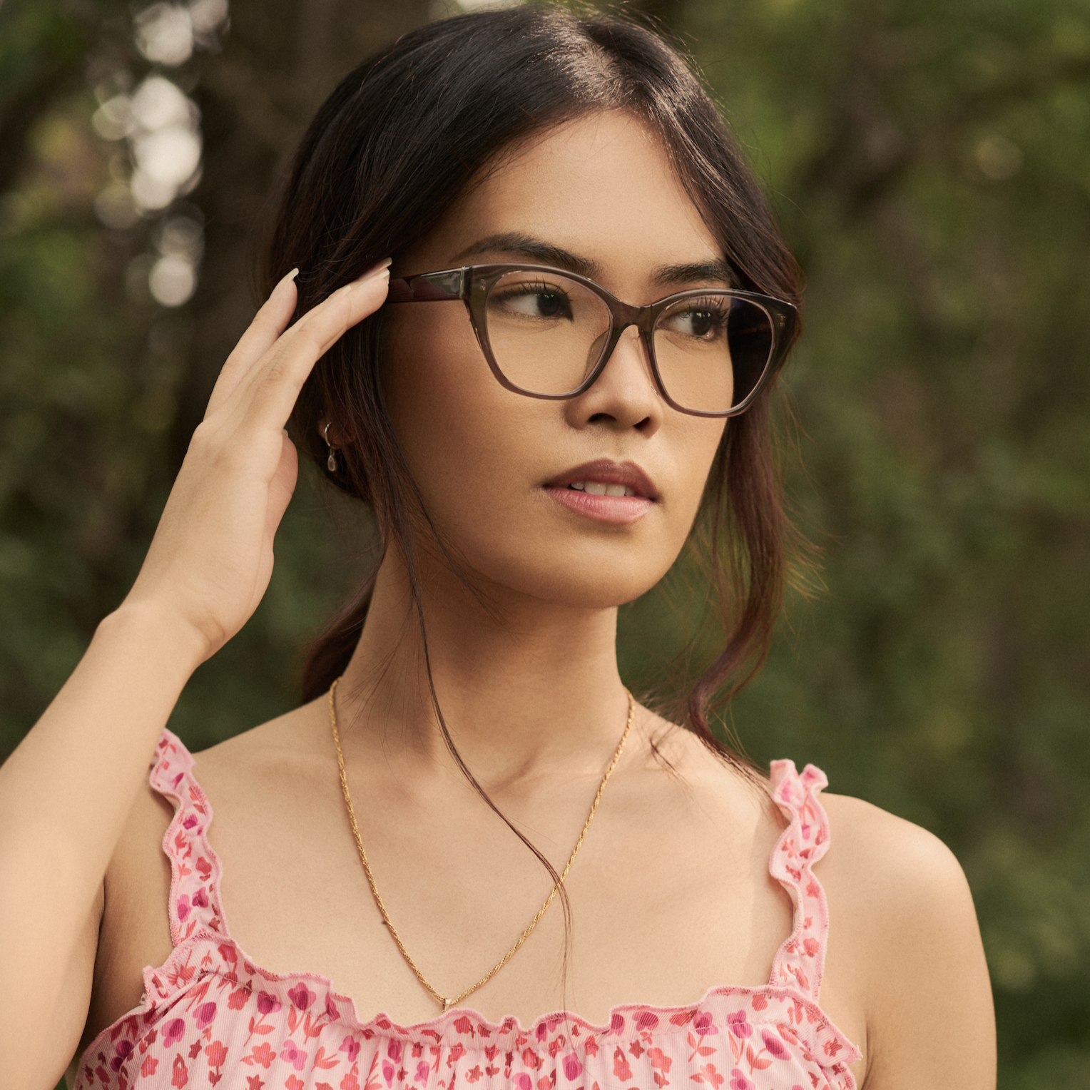 Woman wears KayTran Eyewears Taylor Optical - Deep Taupe glasses and a pink dress, standing outdoors with greenery.