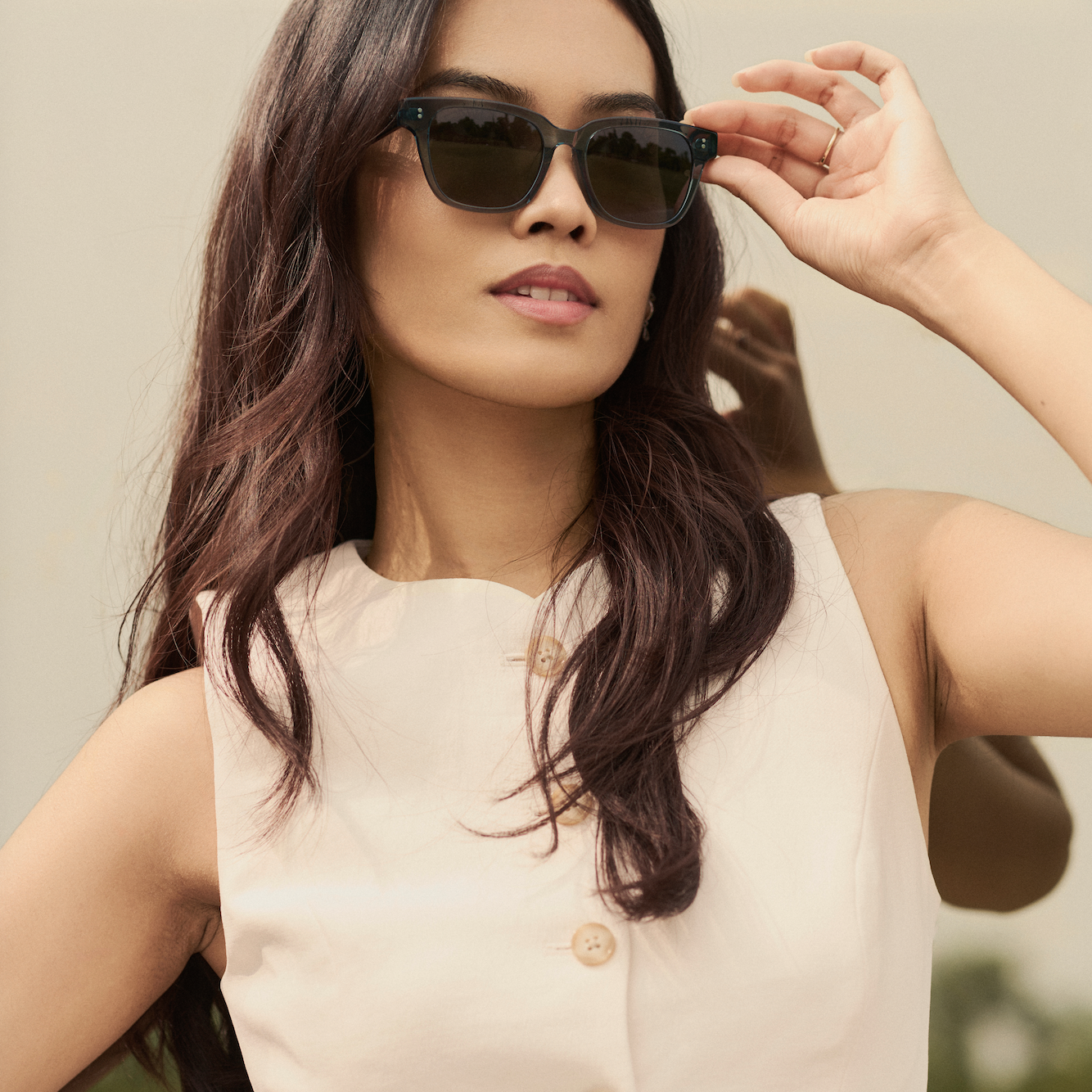Woman with long dark hair wears KayTran Eyewear’s Charlie - Sage sunglasses outdoors, adjusting her glasses.