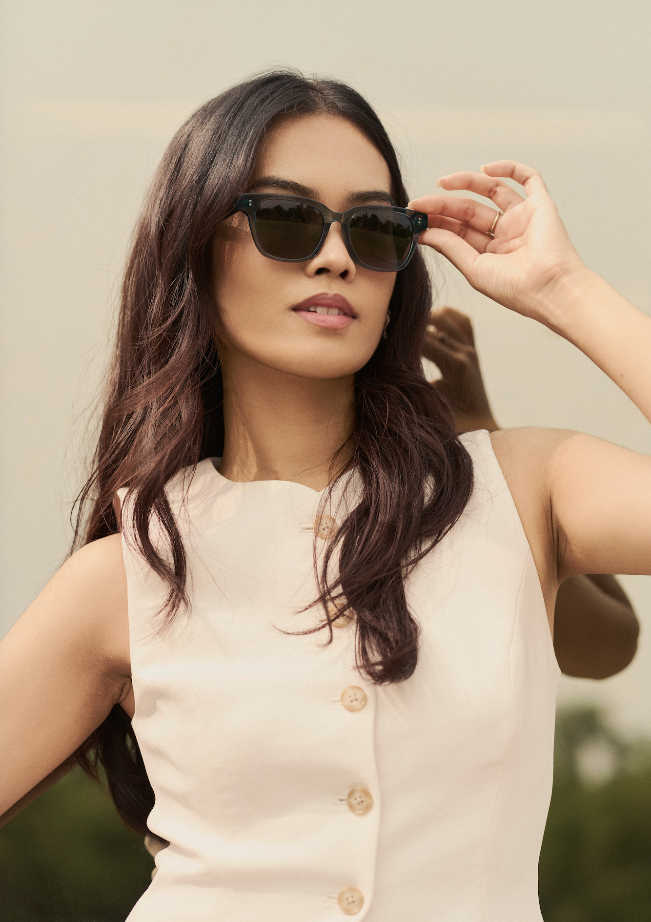 Woman with long dark hair wears KayTran Eyewear’s Charlie - Sage sunglasses outdoors, adjusting her glasses.