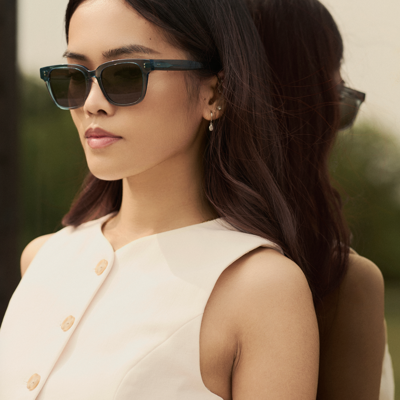 A woman wears KayTran Eyewears Charlie - Sage sunglasses with a sleeveless white top beside a reflective surface.