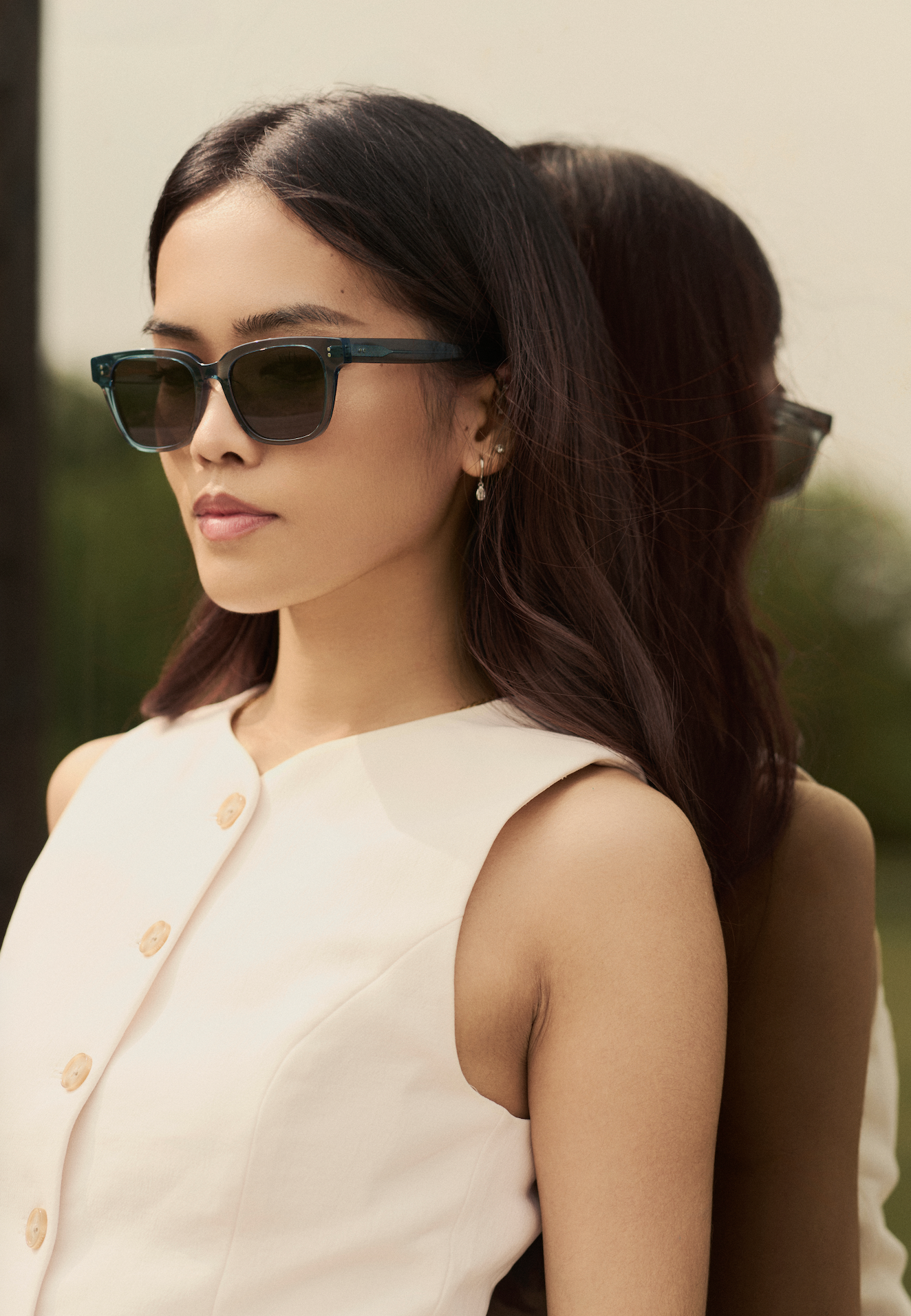 A woman wears KayTran Eyewears Charlie - Sage sunglasses with a sleeveless white top beside a reflective surface.