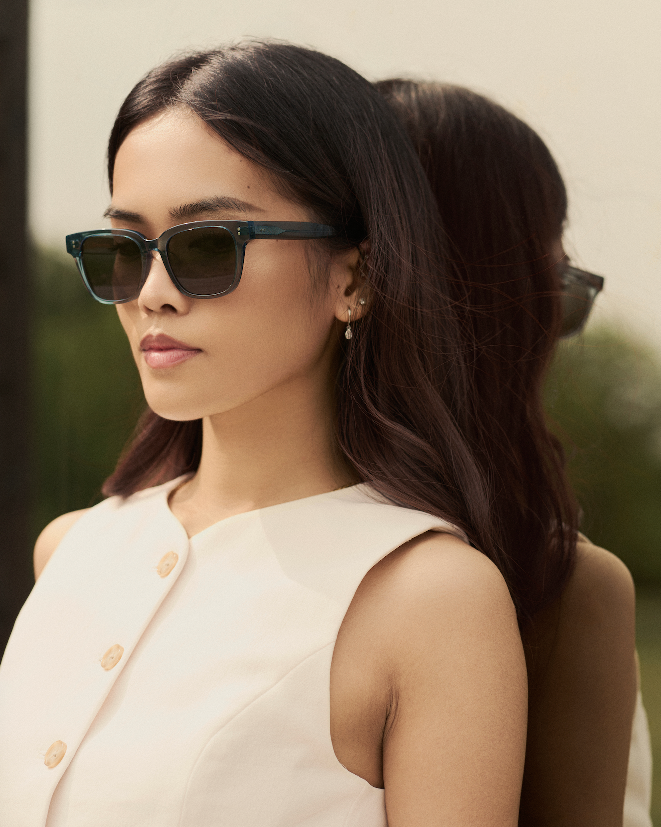 A woman wears KayTran Eyewears Charlie - Sage sunglasses with a sleeveless white top beside a reflective surface.