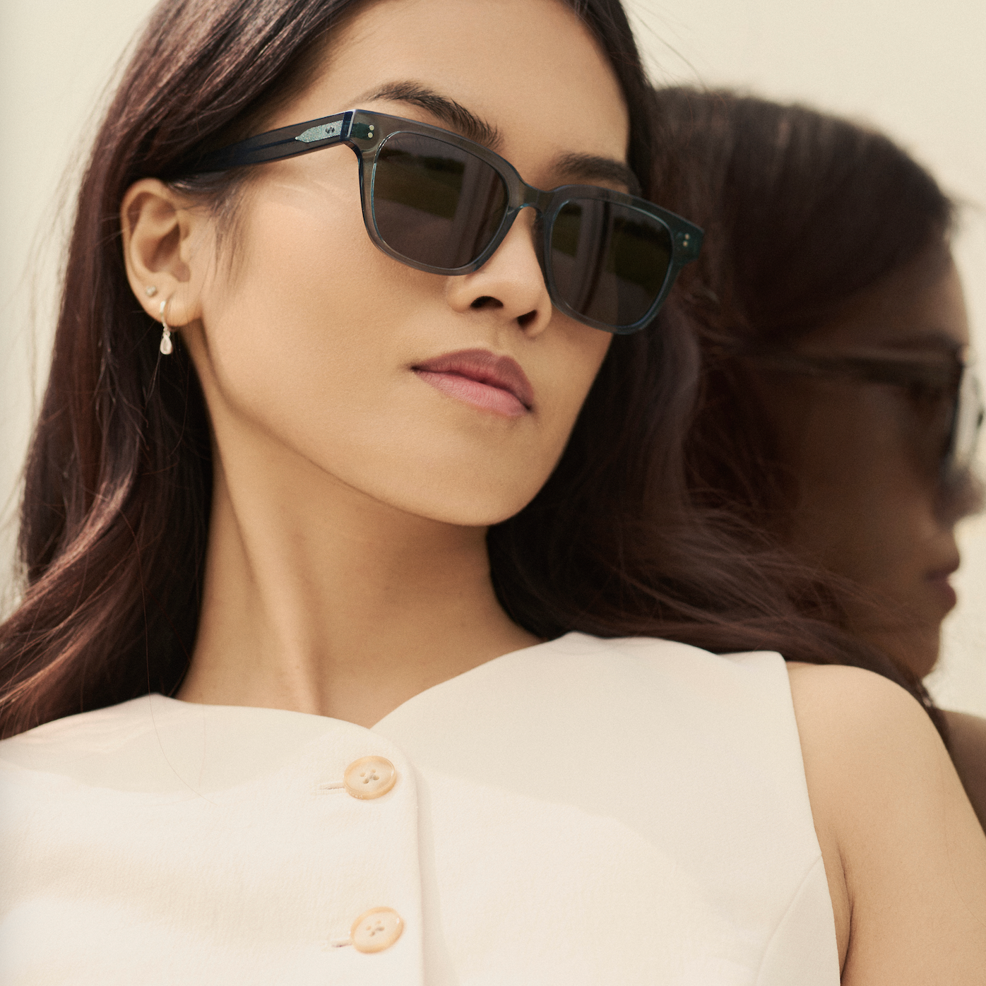 A woman wears KayTran Eyewear’s Charlie - Sage sunglasses and a white top, standing before a mirror.