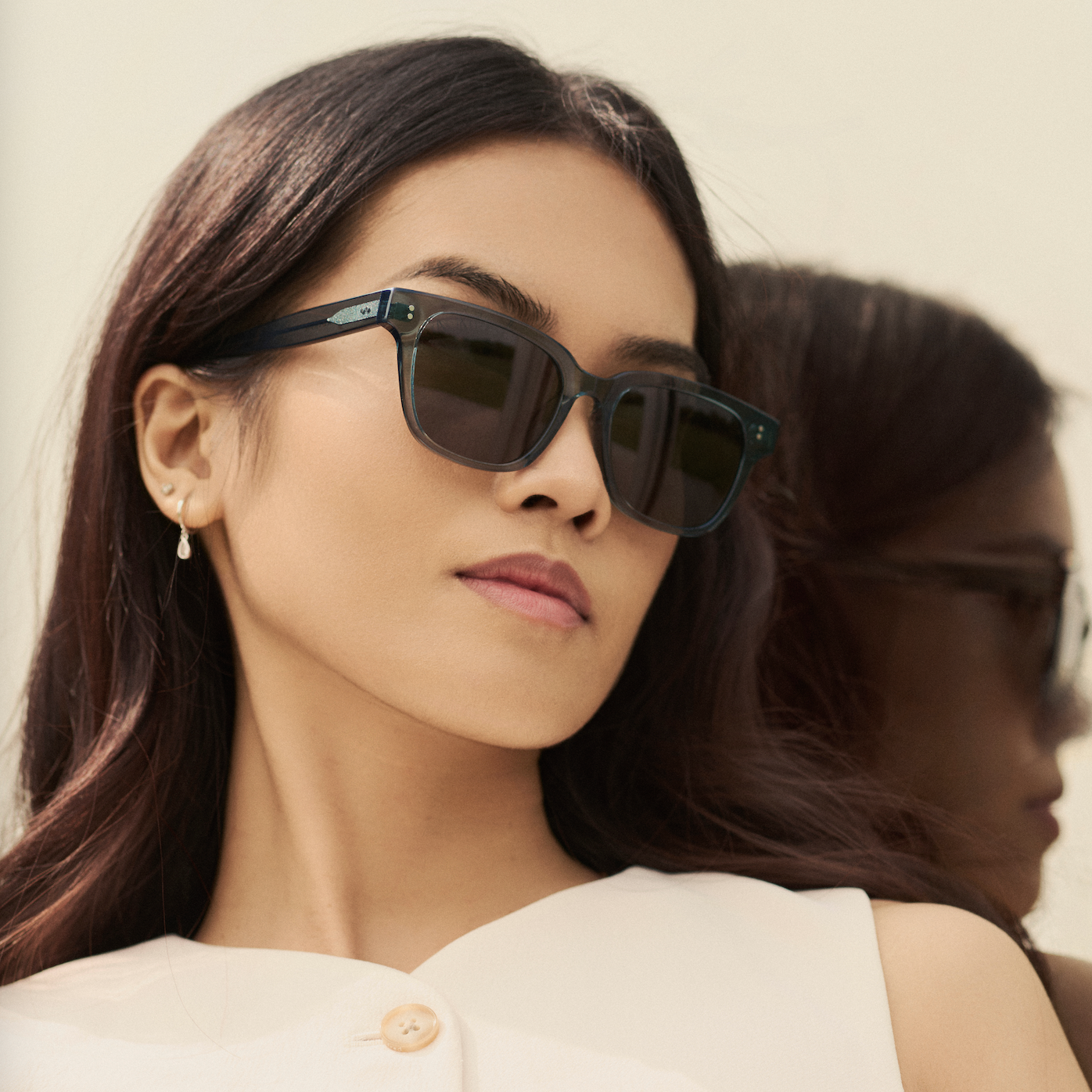 A woman wears KayTran Eyewear’s Charlie - Sage sunglasses and a white top, standing before a mirror.
