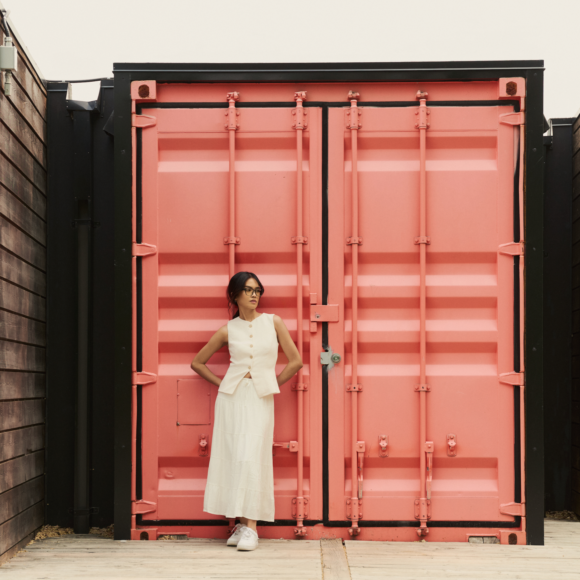 A woman in white wears KayTran Eyewears Charlie Optical - Sage before a pink container, flanked by blue and yellow ones.