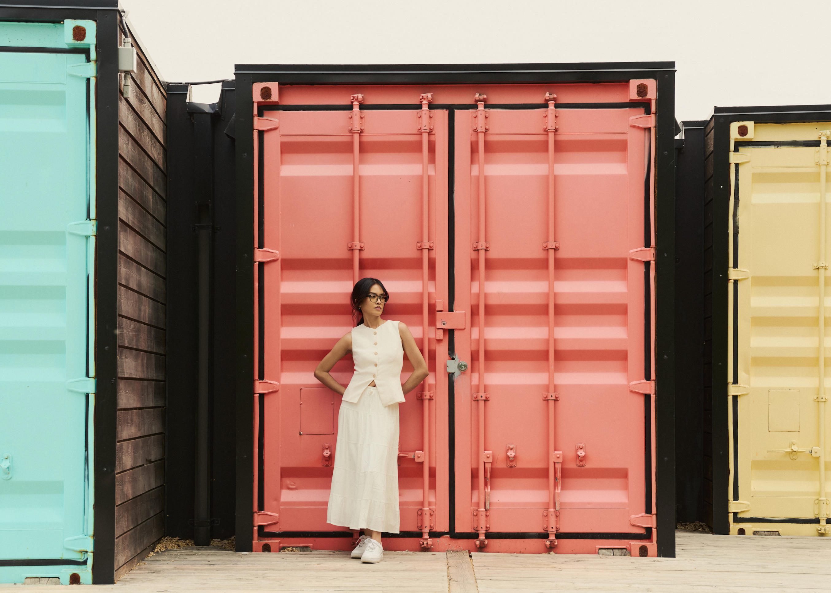 A woman in white wears KayTran Eyewears Charlie Optical - Sage before a pink container, flanked by blue and yellow ones.