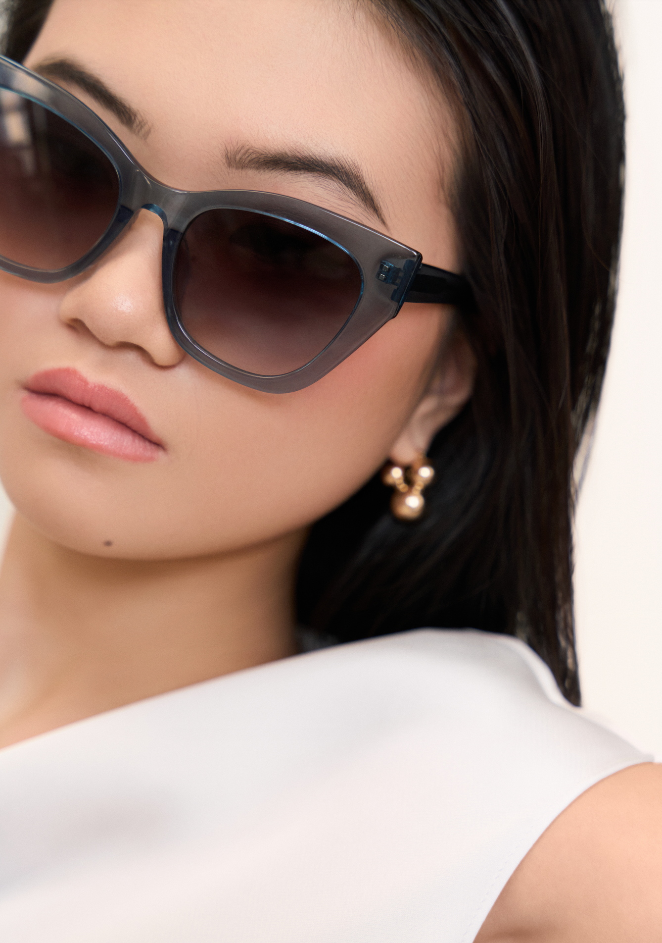 Woman wears KayTran Eyewears Harper - Sea Pine sunglasses, pearl earrings, and white top, looking slightly away.