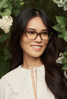 Woman with long dark hair wears KayTran Eyewears Kai Optical - Sage glasses, standing among green leaves and white flowers.