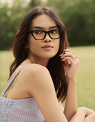 A woman wears KayTran Eyewears Kai Optical - Polished Black glasses outdoors with greenery in the background.
