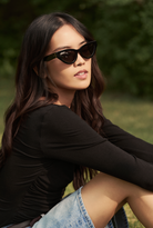 Woman with long dark hair wears KayTran Eyewear Drew - Polished Black sunglasses, black top, and denim skirt outdoors.