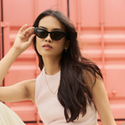 Model wears KayTran Eyewears Drew - Polished Black sunglasses, seated before a pink background, facing the camera.