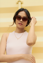 Woman wears KayTran Eyewear Drew - Burgundy sunglasses and a white sleeveless top before a yellow striped background.
