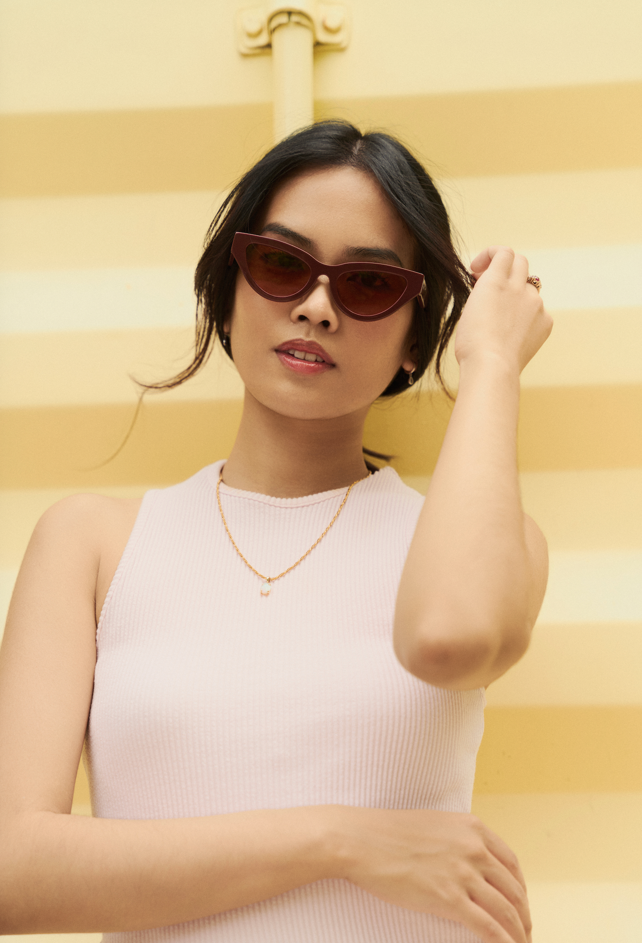 Woman wears KayTran Eyewear Drew - Burgundy sunglasses and a white sleeveless top before a yellow striped background.