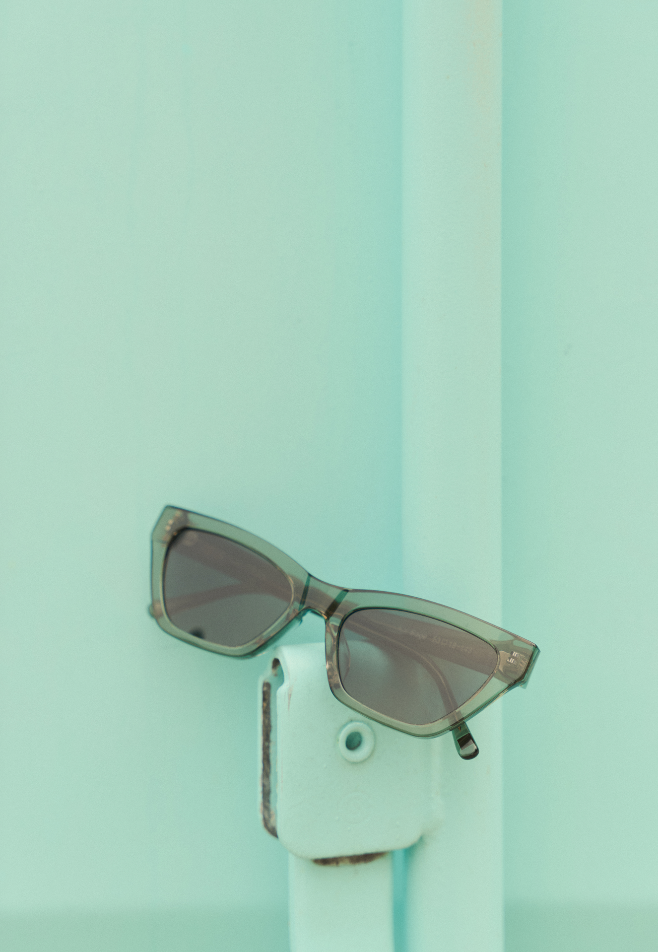 The Kai - Sage sunglasses by KayTran Eyewear rest on a light teal metal fixture against a matching teal background.