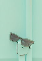 The Kai - Sage sunglasses by KayTran Eyewear rest on a light teal metal fixture against a matching teal background.