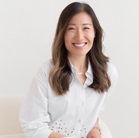 Woman with long brown hair sits and smiles, wearing a white button-up shirt against a plain light background.