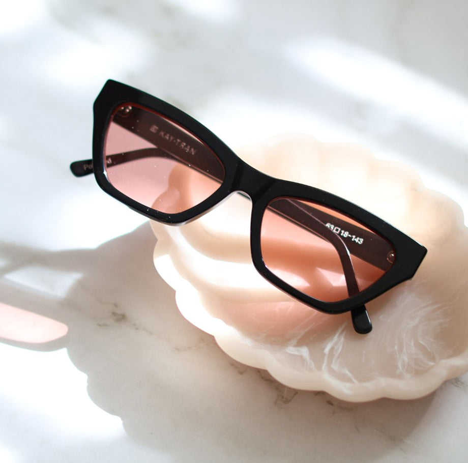 Black-framed sunglasses with pink lenses rest on a shell-shaped dish on a white marble surface.