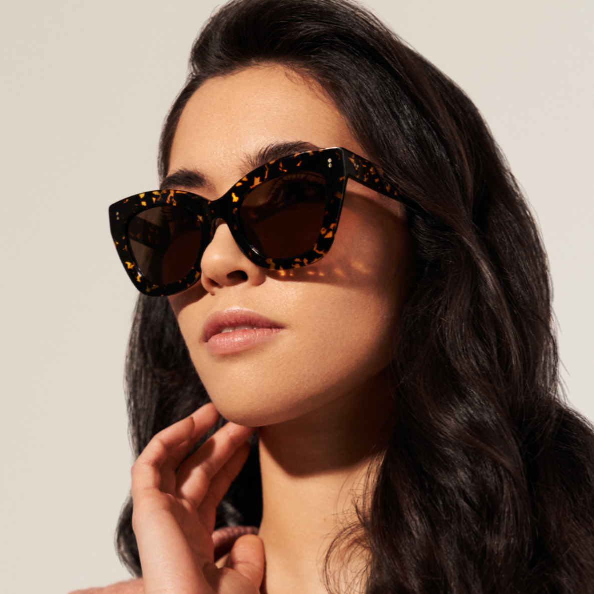 Woman with long dark hair wears KayTran Eyewear Arlo - Amber Scale Tort cat-eye sunglasses and a light pink top.