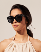 A woman in KayTran Eyewears Rowan - Polished Black sunglasses and a light halter top looks ahead.