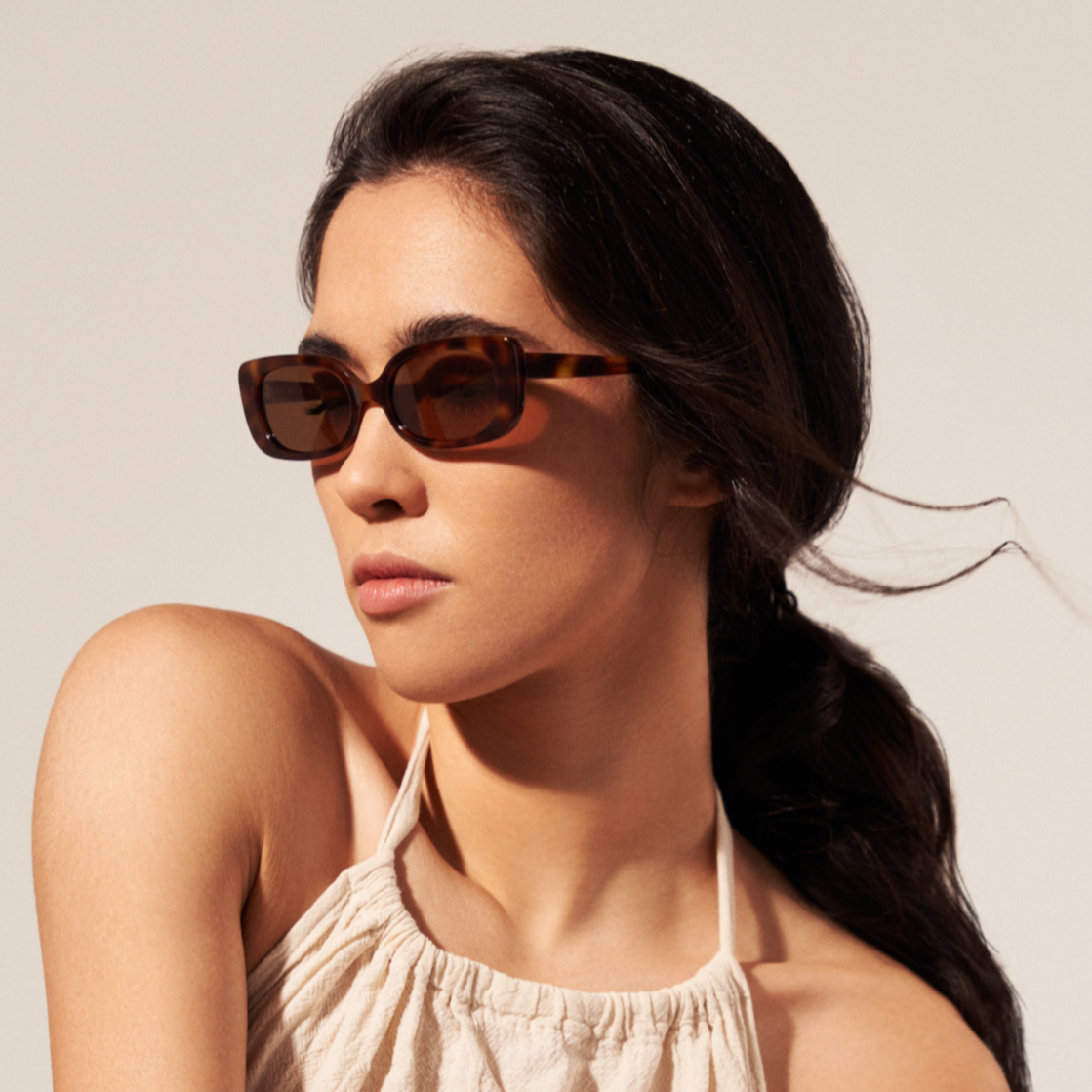 Woman with long dark hair wearing KayTran Eyewears Billie - Lava Tortoise sunglasses and a cream halter top, looking sideways.