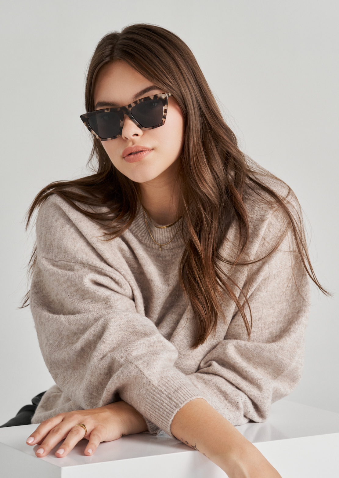 Woman in beige sweater wears KayTran Eyewears Frankie - Chocolate Chip sunglasses at a white table, plain background.