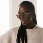 A woman with braided hair wears KayTran Eyewears Charlie - Stone Taupe sunglasses and a beige sweater, looking down.