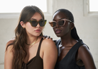 Two women wear KayTran Eyewears Harper - Almond sunglasses indoors, standing close and looking off camera.