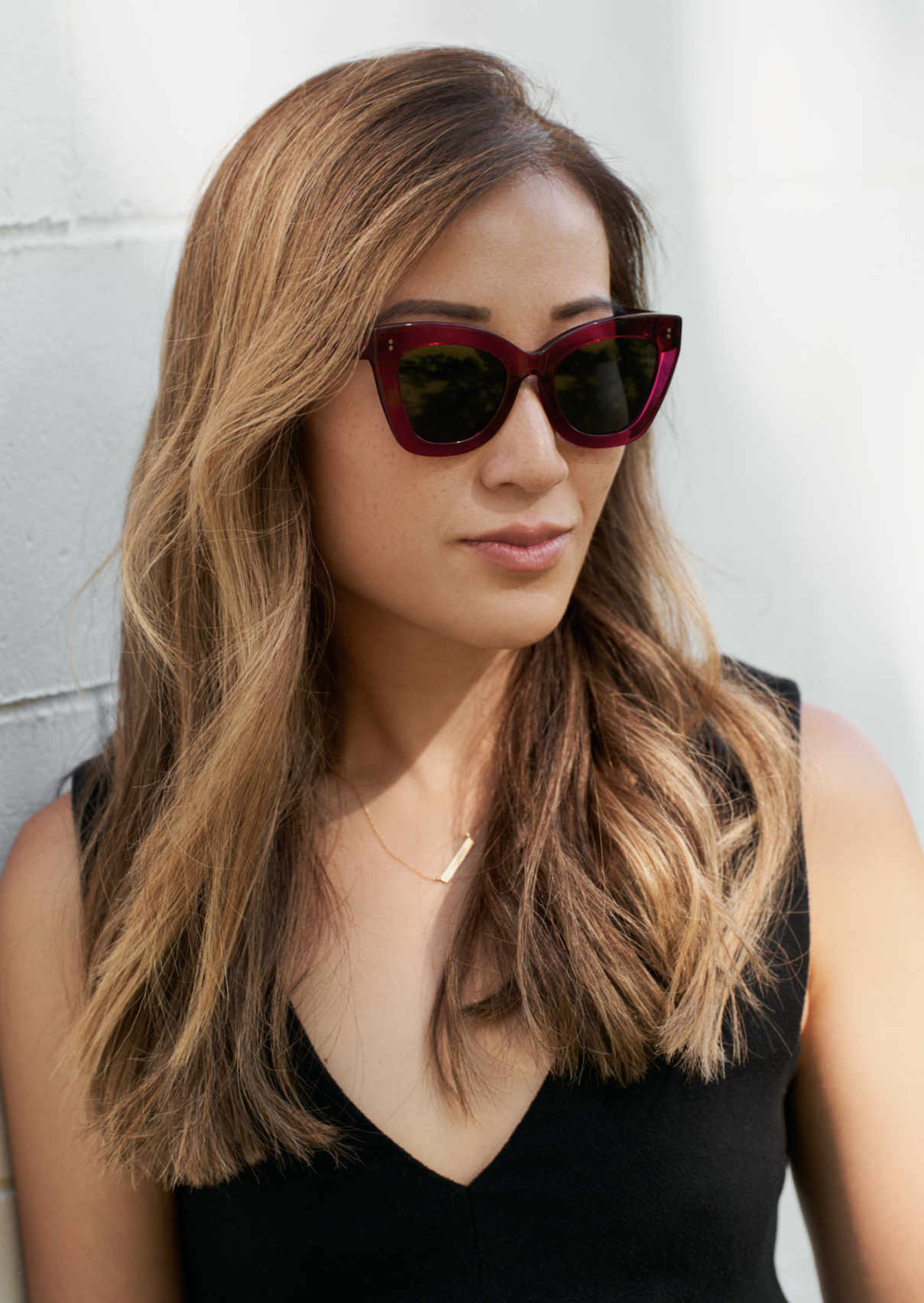 Woman with long, light brown hair wears KayTran Eyewear Arlo - Berry sunglasses and a black top against a white wall.