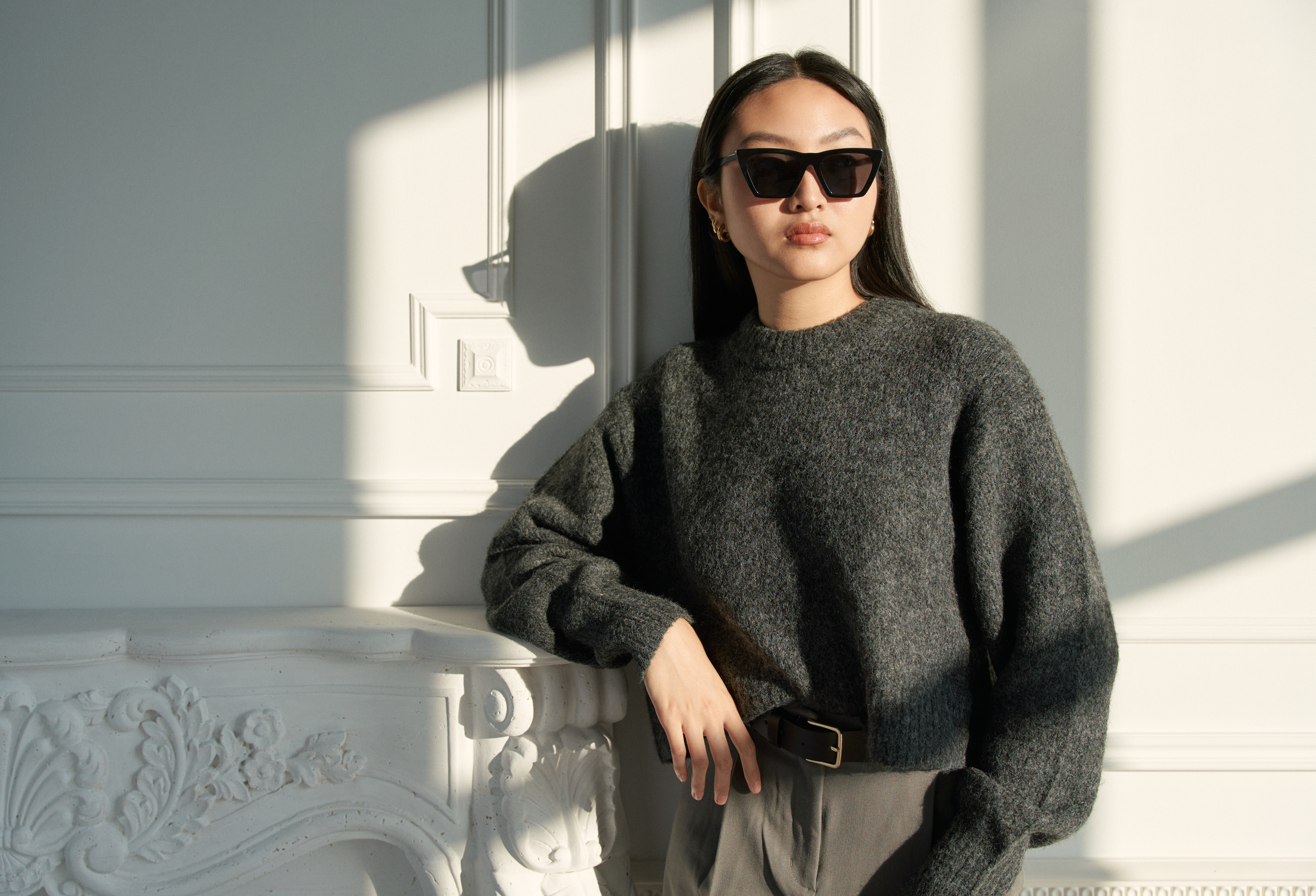 Woman wears KayTran Eyewears Frankie - Polished Black sunglasses, leaning by a white mantel in a sunlit room.