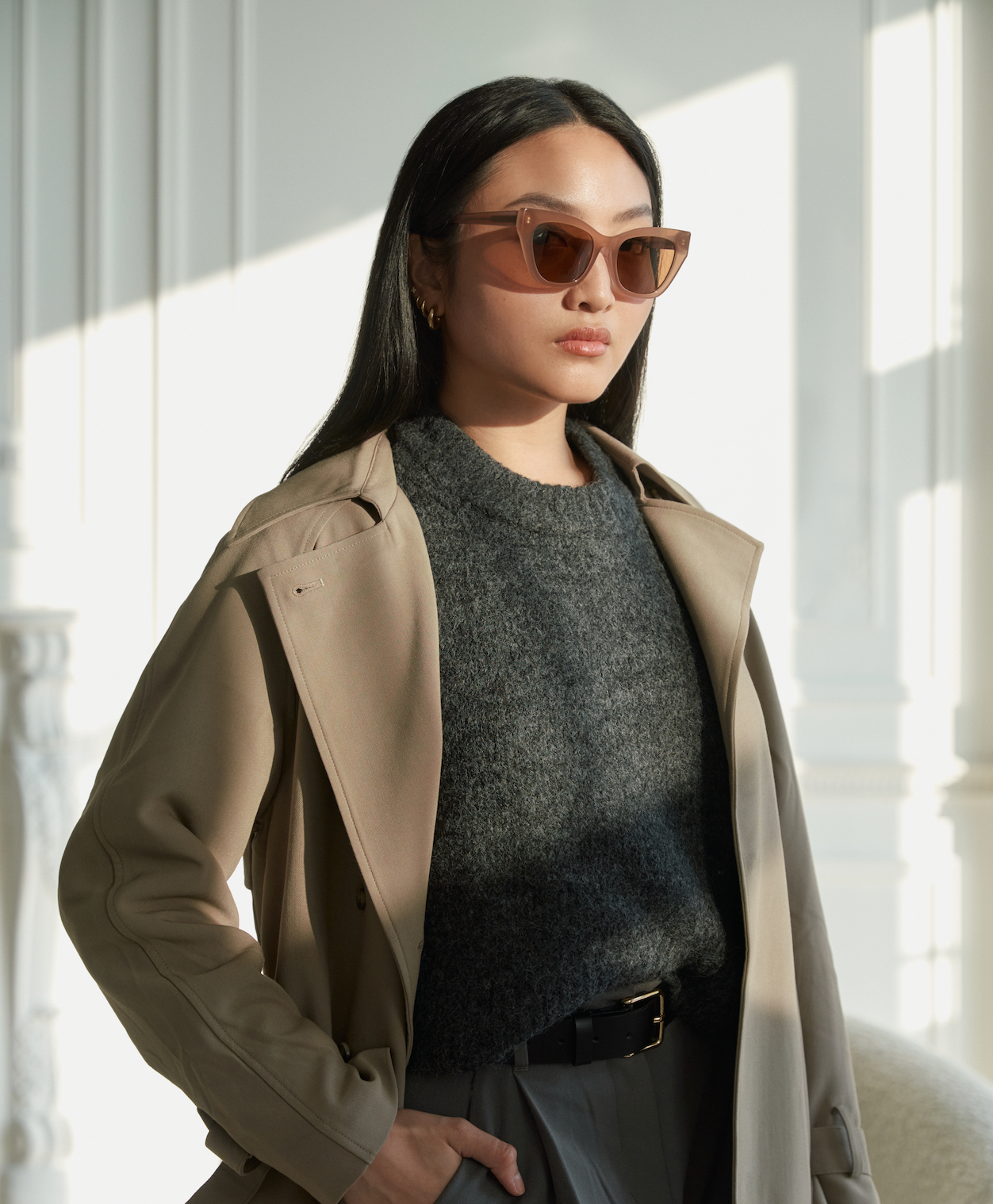 A woman wears KayTran Eyewears Harper - Almond sunglasses, a beige coat, and a dark sweater indoors with sunlight.