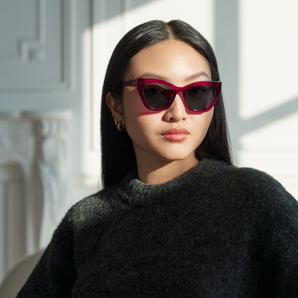 Woman with long dark hair wears KayTran Eyewear Harper - Berry sunglasses and a dark sweater in a sunlit room.