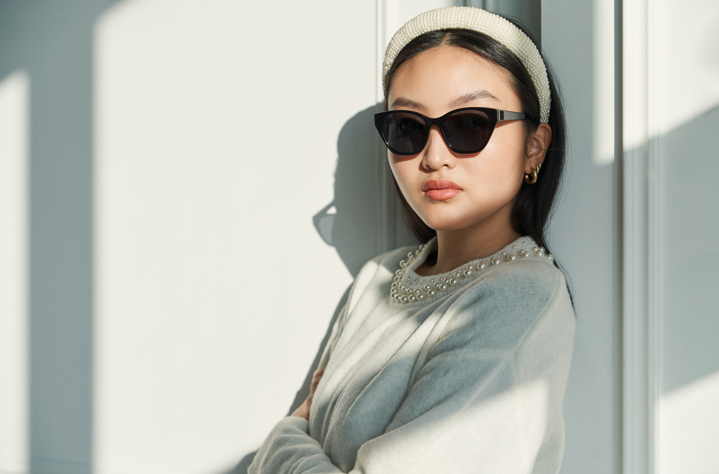 A woman in KayTran Eyewears Taylor - Polished Black sunglasses stands by a white wall in natural light.