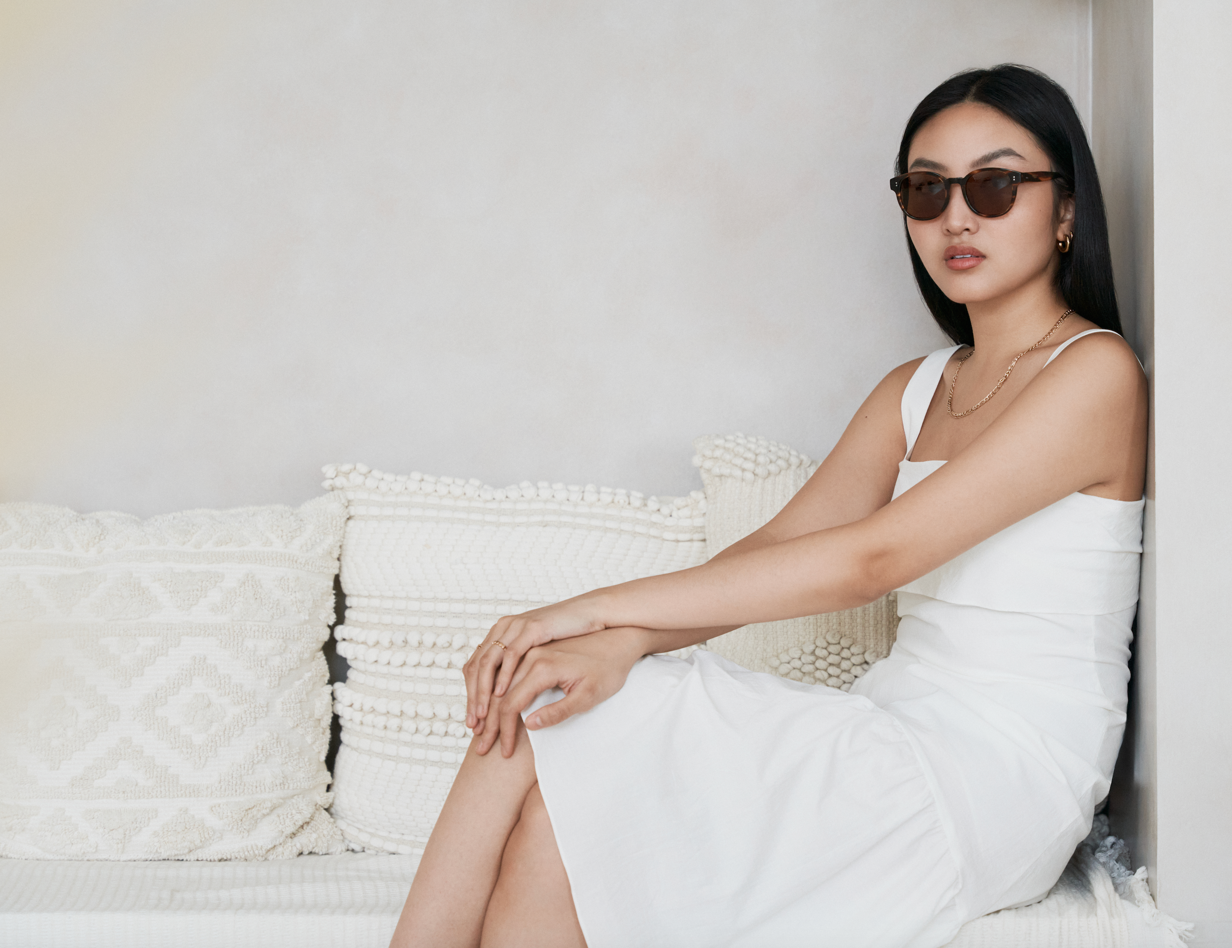 Woman in a white dress wears KayTran Eyewears Reese - Amber Honey sunglasses on a bench with textured pillows.