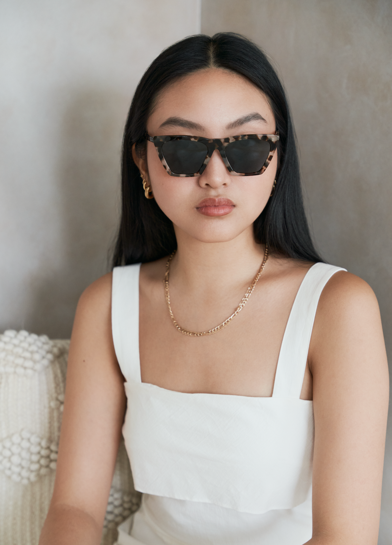 Woman with long dark hair wearing KayTran Eyewear Frankie - Chocolate Chip sunglasses, white sleeveless top, and gold jewelry.