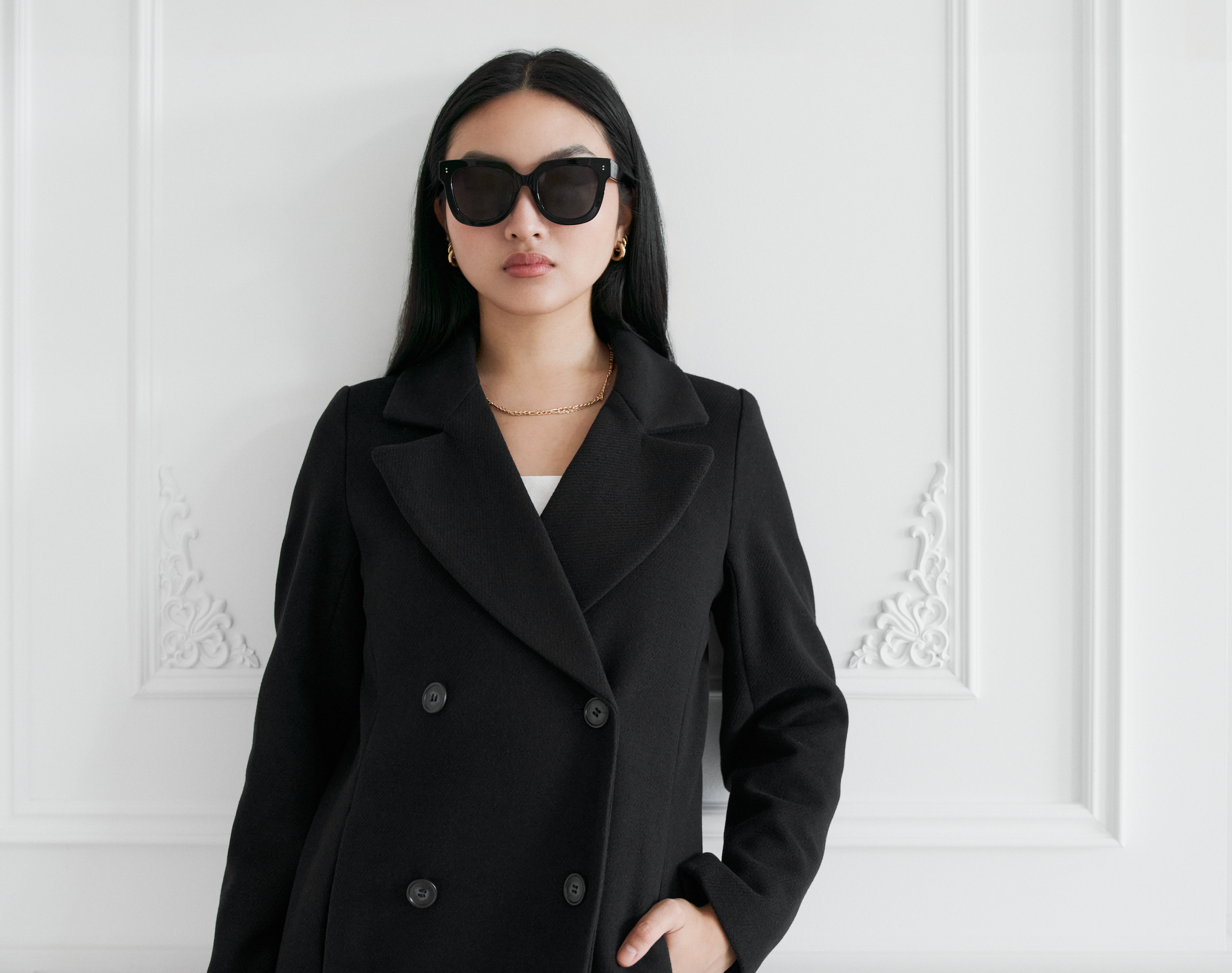 Woman wears KayTran Eyewears Rowan - Polished Black sunglasses and stands by an ornate white wall.