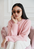 Woman with long black hair wears KayTran Eyewear Arlo - Milky Blush sunglasses and a pink sweater, seated on a pink chair.
