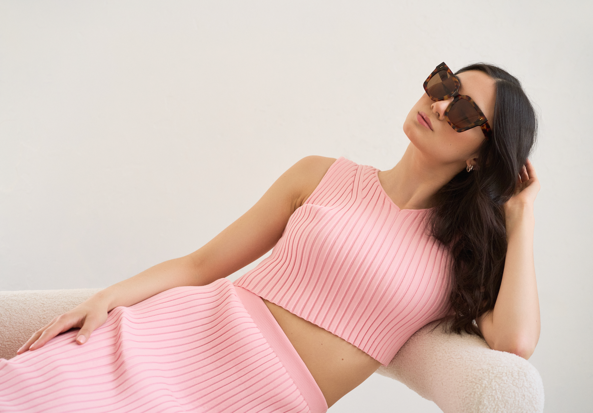 Woman in a ribbed pink crop top and skirt reclines on a chair, wearing KayTran Eyewear’s Logan - Ember & Brown Lens.