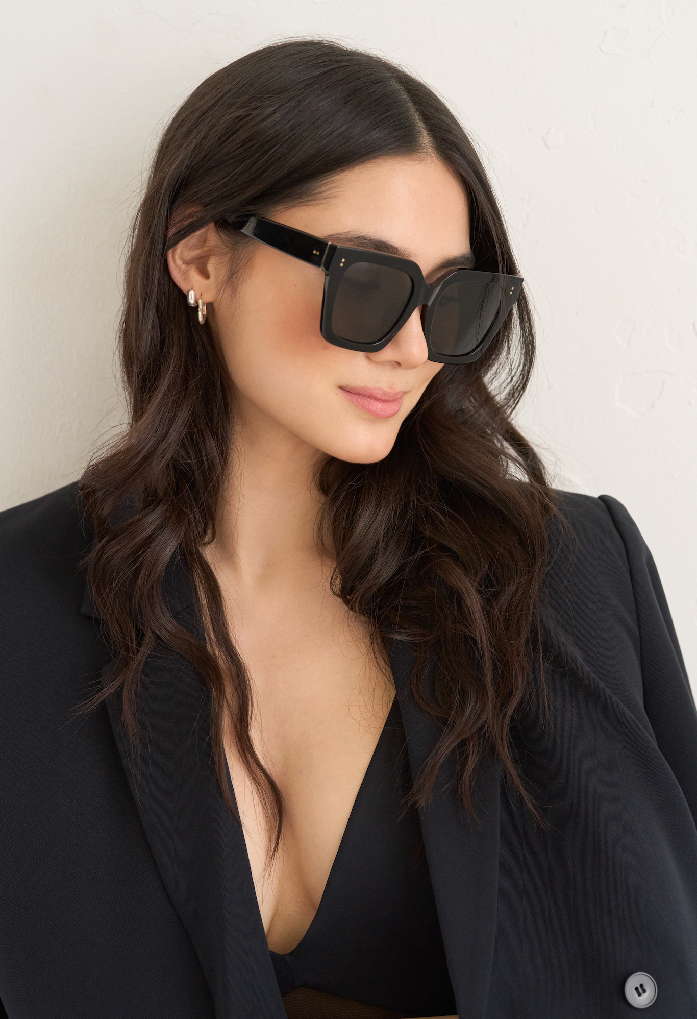 Woman with long wavy hair wears KayTran Eyewear’s Logan - Polished Black sunglasses, black blazer, and top.