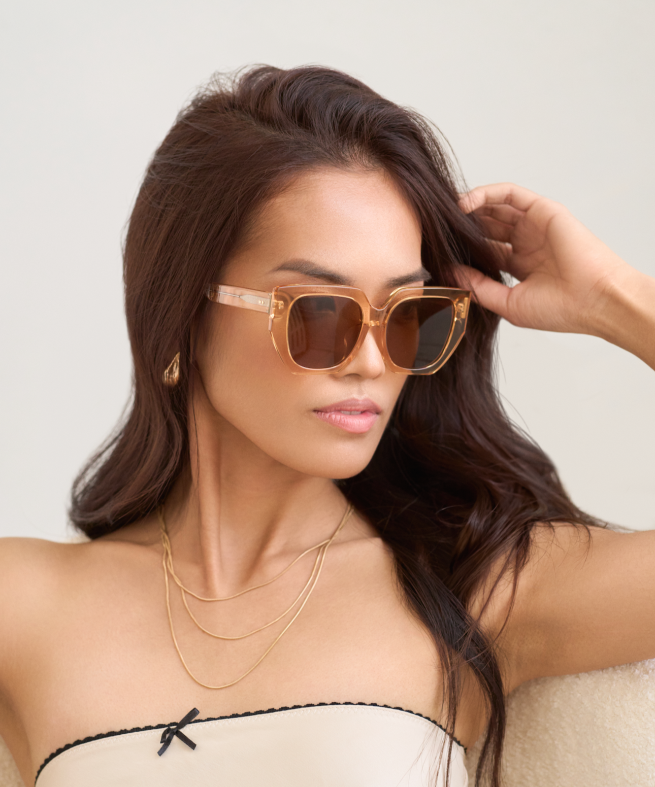 Woman with long dark hair wears KayTran Eyewear’s Luna - Apricot sunglasses, gold necklaces, and a strapless top.