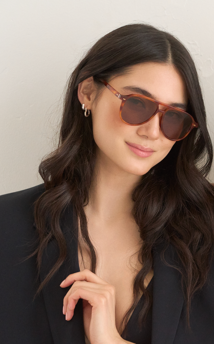Woman with long dark hair wears KayTran Eyewears Knox - Cider sunglasses, a black blazer, and small hoop earrings.
