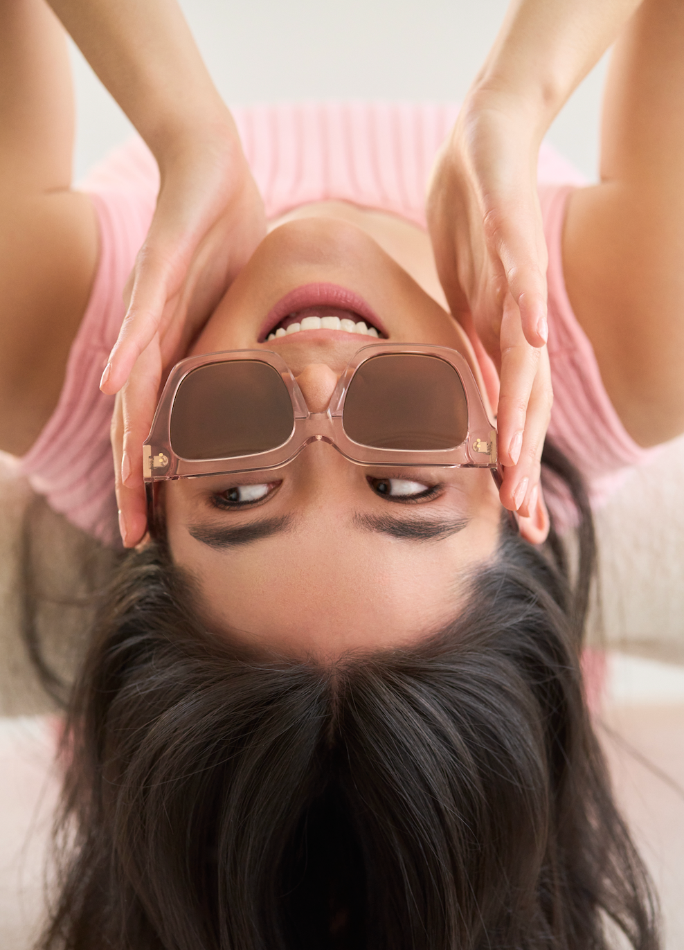 A woman lies upside down, smiling slightly while holding KayTran Eyewears Logan - Bloom square sunglasses over her eyes.
