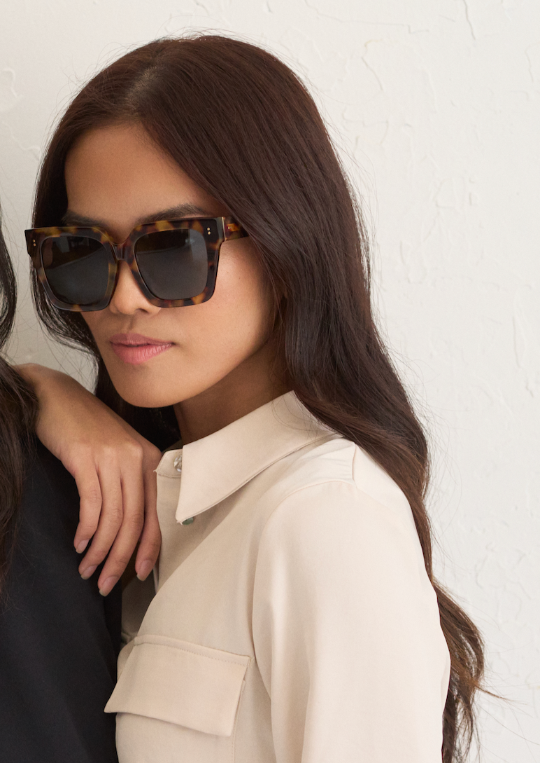 Woman with long brown hair wears KayTran Eyewears Logan - Ember & Grey Lens sunglasses, beige shirt, white wall behind.