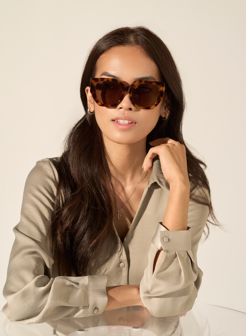 Woman with long dark hair wears KayTran Eyewear’s Luna - Honey Tort sunglasses and a beige blouse at a glass table.